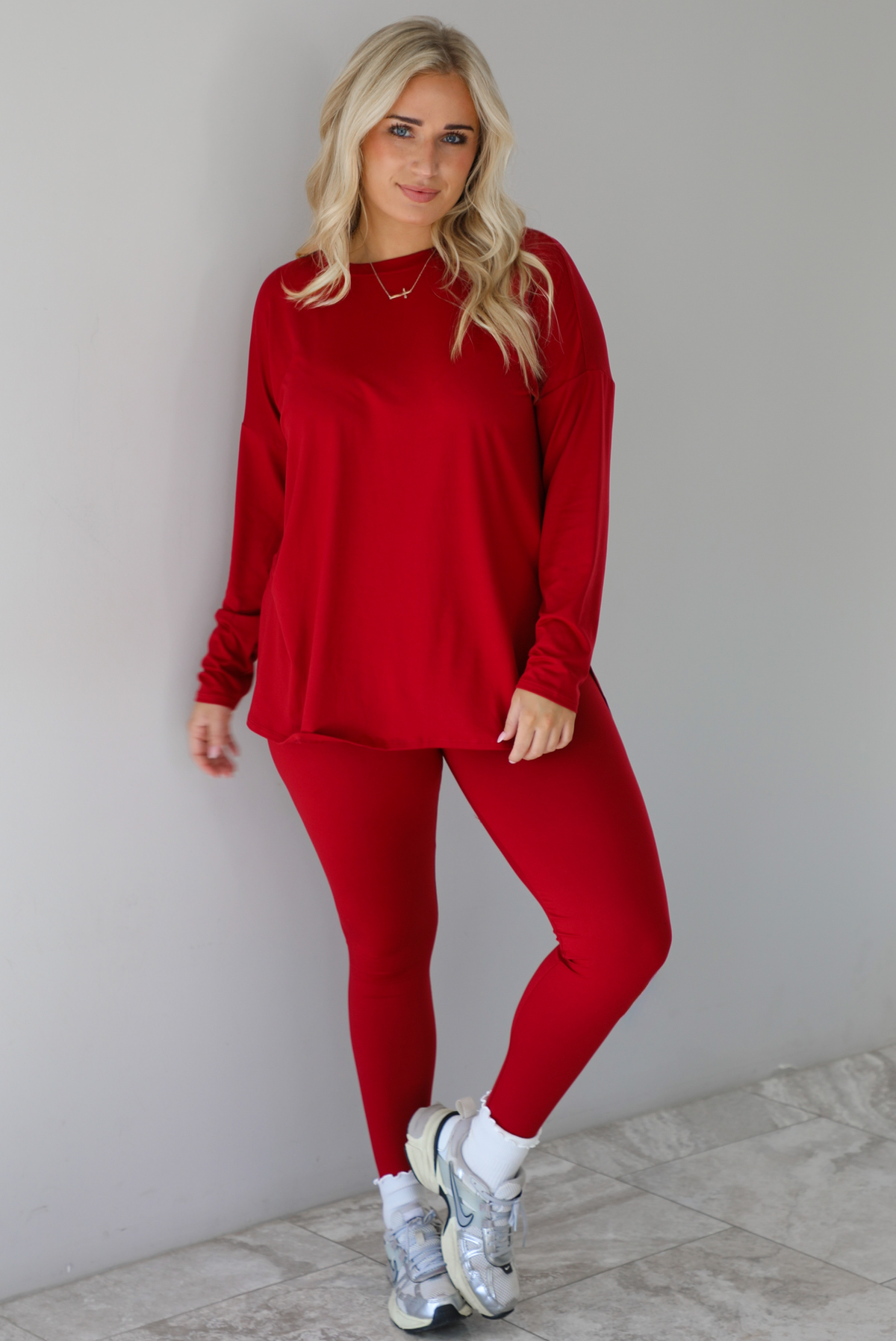 Woman wearing a red two piece set that features an oversized long sleeved top and fitted leggings. The set is paired with high ankle socks and silver sneakers. The photo is taken indoors against a white wall with white marble flooring.