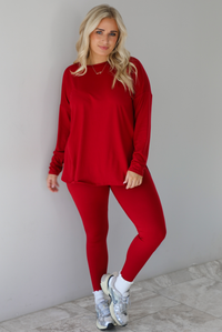 Woman wearing a red two piece set that features an oversized long sleeved top and fitted leggings. The set is paired with high ankle socks and silver sneakers. The photo is taken indoors against a white wall with white marble flooring.