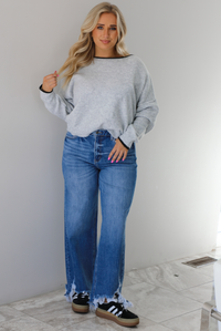 Woman wearing a grey relaxed fit sweater that has black trim on it, medium wash distressed denim jeans, and black sneakers. The photo is taken indoors against a white wall with white marble flooring. 