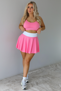 Woman wearing a pink and white two piece set featuring a thin strapped sports bra and high waisted skirt. The set is paired with high white ankle socks and silver sneakers. The photo is taken indoors against a white wall with white marble flooring. 