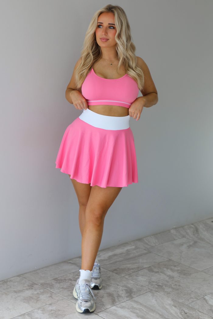 Woman wearing a pink and white two piece set featuring a thin strapped sports bra and high waisted skirt. The set is paired with high white ankle socks and silver sneakers. The photo is taken indoors against a white wall with white marble flooring. 
