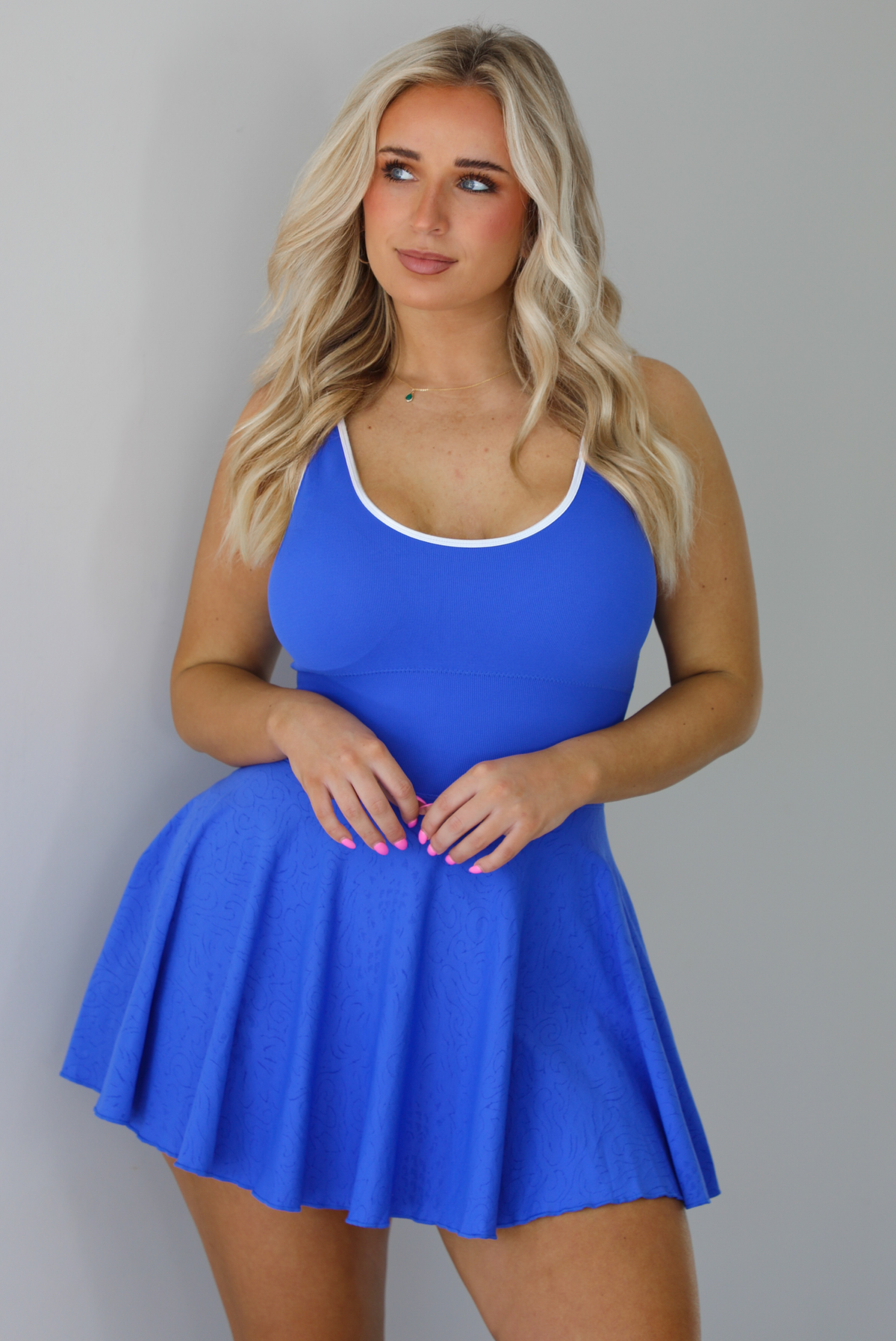 Woman wearing a blue athleisure dress that has white trim, a fitted bodice, and flowy skirt. The photo is taken indoors against a white wall.