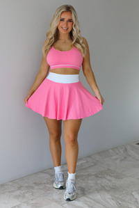 Woman wearing a pink and white two piece set featuring a thin strapped sports bra and high waisted skirt. The set is paired with high white ankle socks and silver sneakers. The photo is taken indoors against a white wall with white marble flooring. 