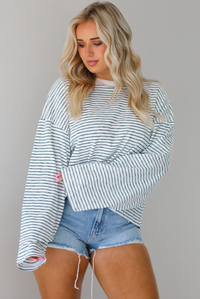 Woman wearing an oversized white and black striped, long sleeved, box tee, with a light wash denim shorts. The top is tucked into the front of the shorts. The photo is taken indoors against a white wall.