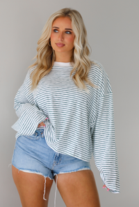 Woman wearing an oversized white and black striped, long sleeved, box tee, with a light wash denim shorts. The photo is taken indoors against a white wall.