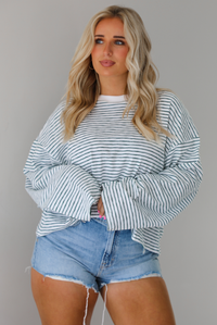 Woman wearing an oversized white and black striped, long sleeved, box tee, with a light wash denim shorts. The top is tucked into the front of the shorts. The photo is taken indoors against a white wall.