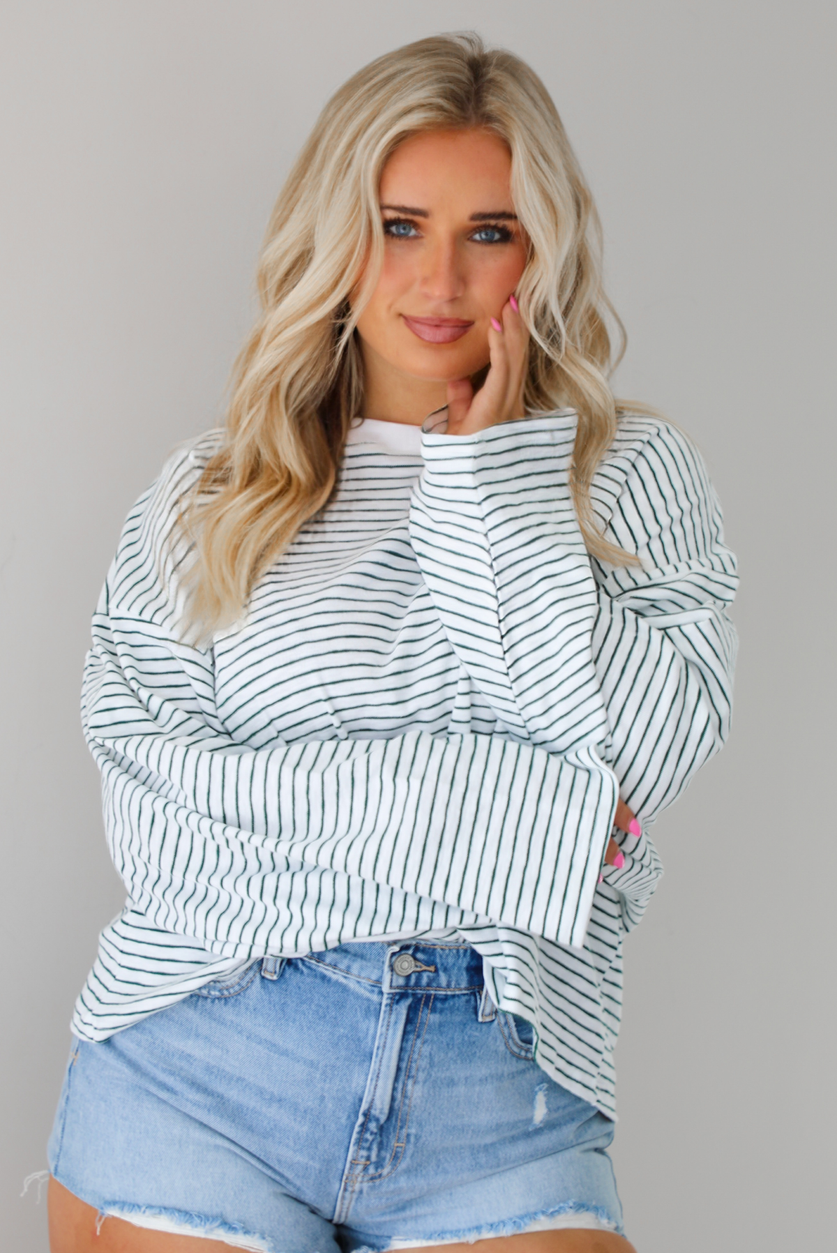 Woman wearing an oversized white and black striped, long sleeved, box tee, with a light wash denim shorts. The top is tucked into the front of the shorts. The photo is taken indoors against a white wall.