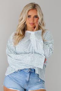 Woman wearing an oversized white and black striped, long sleeved, box tee, with a light wash denim shorts. The top is tucked into the front of the shorts. The photo is taken indoors against a white wall.