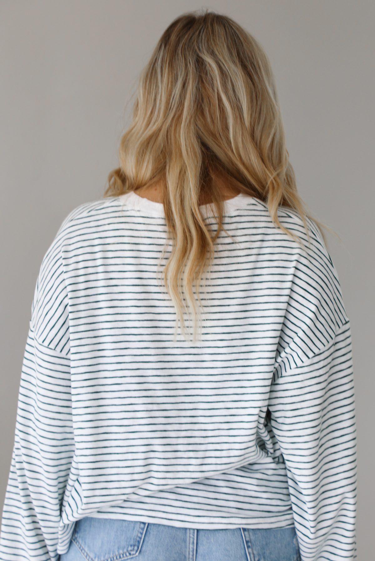 Woman wearing an oversized white and black striped, long sleeved, box tee, with a light wash denim shorts. The photo shows the back of the top. The photo is taken indoors against a white wall.