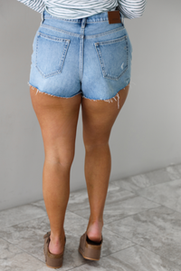Woman wearing a light-wash pair of denim shorts that have distressed detailing. The photo shows the back of the shorts. The photo is taken indoors against a white wall with white marble flooring. 