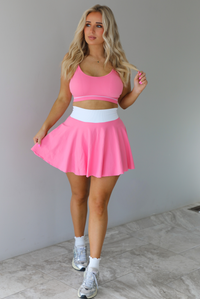 Woman wearing a pink and white two piece set featuring a thin strapped sports bra and high waisted skirt. The set is paired with high white ankle socks and silver sneakers. The photo is taken indoors against a white wall with white marble flooring. 