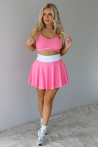 Woman wearing a pink and white two piece set featuring a thin strapped sports bra and high waisted skirt. The set is paired with high white ankle socks and silver sneakers. The photo is taken indoors against a white wall with white marble flooring. 