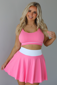 Woman wearing a pink and white two piece set featuring a thin strapped sports bra and high waisted skirt.  The photo is taken indoors against a white wall.