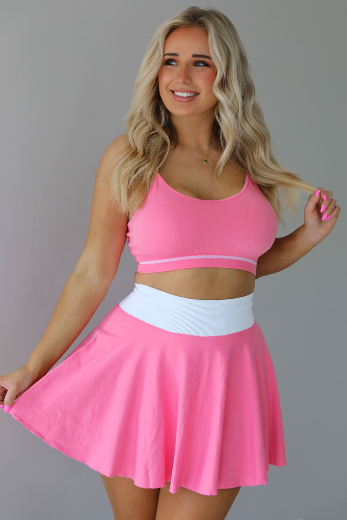Woman wearing a pink and white two piece set featuring a thin strapped sports bra and high waisted skirt.  The photo is taken indoors against a white wall.