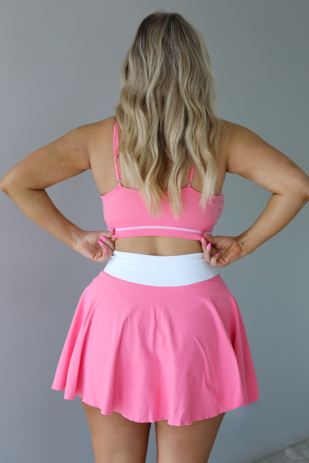 Woman wearing a pink and white two piece set featuring a thin strapped sports bra and high waisted skirt.  The photo is taken indoors against a white wall. The photo shows the back of the set. 