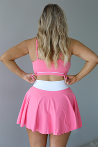 Woman wearing a pink and white two piece set featuring a thin strapped sports bra and high waisted skirt.  The photo is taken indoors against a white wall. The photo shows the back of the set. 
