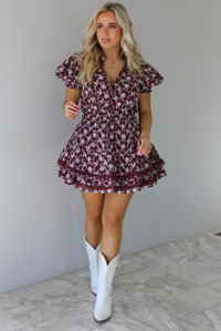 Woman wearing a wine dress that has an embroidered white floral print pattern, v-neckline, puffed sleeves, cinched with elastic waistline, and puffed hem skirt. The dress is paired with white cowboy boots. The photo is taken indoors against a white wall with white marble flooring.