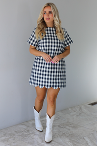 Woman wearing a dress with black and white gingham print pattern throughout. The dress has short sleeves, a high neckline, relaxed fit design, functional pockets on either side, and a scalloped hem. The dress is paired with white cowboy boots. The photo is taken in front of a white wall with white marble flooring. 