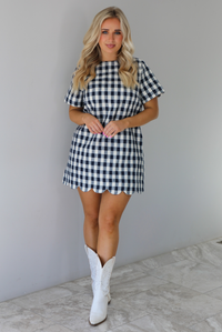 Woman wearing a dress with black and white gingham print pattern throughout. The dress has short sleeves, a high neckline, relaxed fit design, functional pockets on either side, and a scalloped hem. The dress is paired with white cowboy boots. The photo is taken in front of a white wall with white marble flooring. 