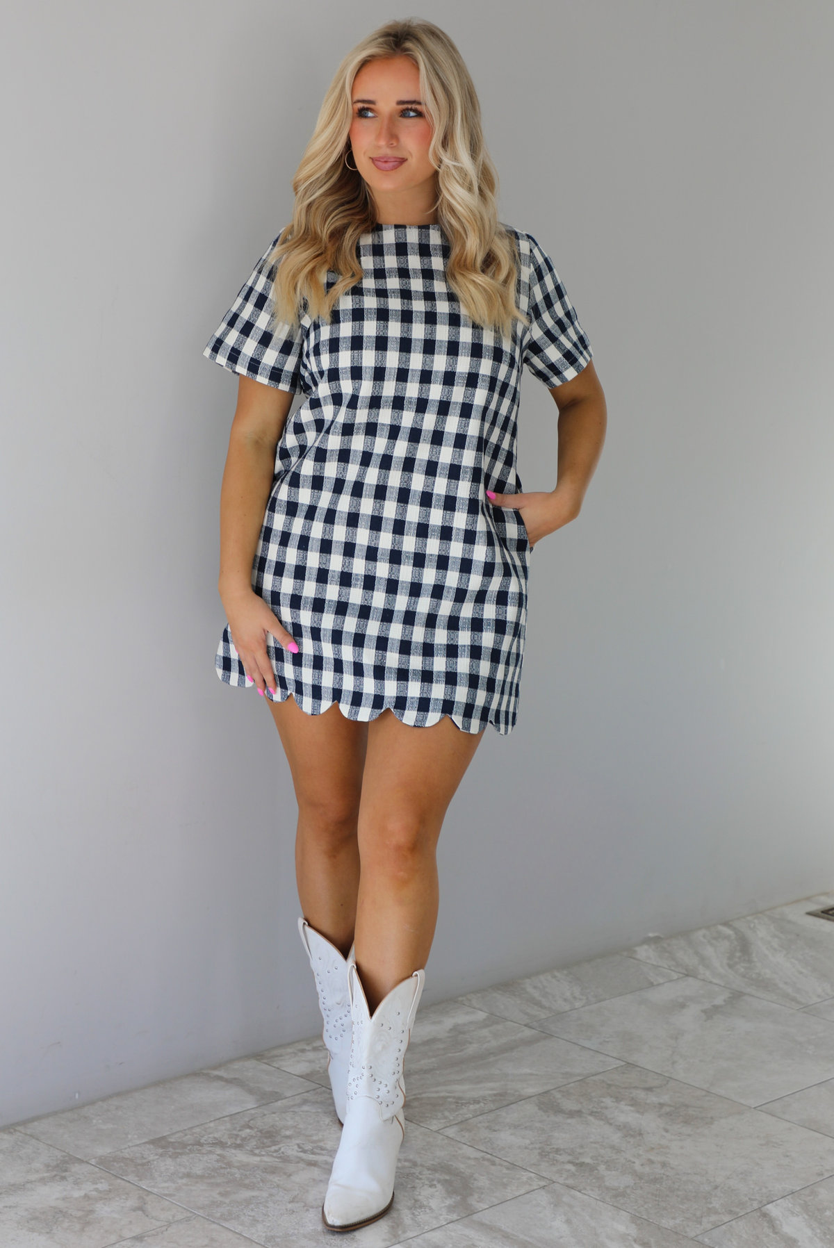 Woman wearing a dress with black and white gingham print pattern throughout. The dress has short sleeves, a high neckline, relaxed fit design, functional pockets on either side, and a scalloped hem. The dress is paired with white cowboy boots. The photo is taken in front of a white wall with white marble flooring. 