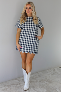 Woman wearing a dress with black and white gingham print pattern throughout. The dress has short sleeves, a high neckline, relaxed fit design, functional pockets on either side, and a scalloped hem. The dress is paired with white cowboy boots. The photo is taken in front of a white wall with white marble flooring. 