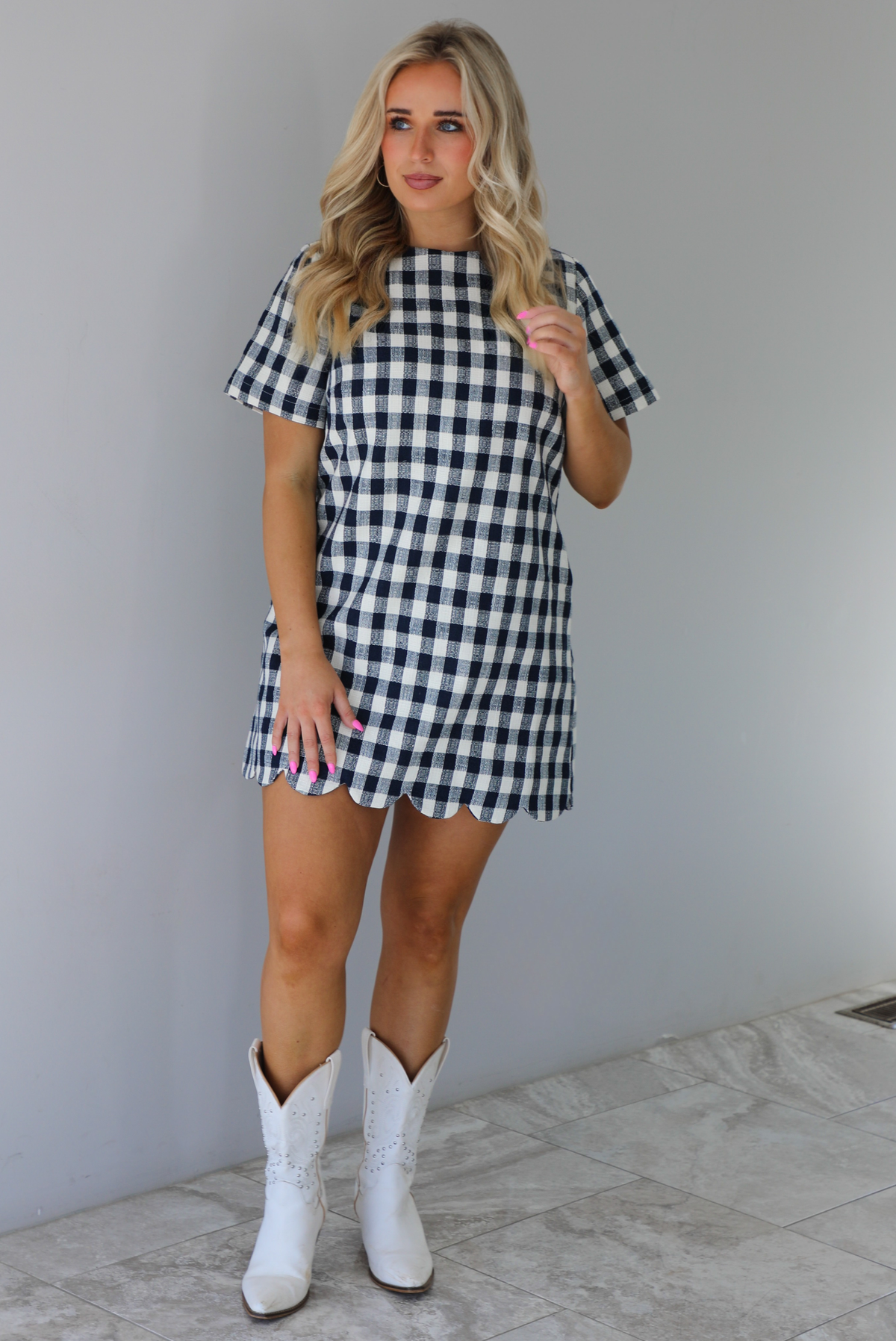 Woman wearing a dress with black and white gingham print pattern throughout. The dress has short sleeves, a high neckline, relaxed fit design, functional pockets on either side, and a scalloped hem. The dress is paired with white cowboy boots. The photo is taken in front of a white wall with white marble flooring. 