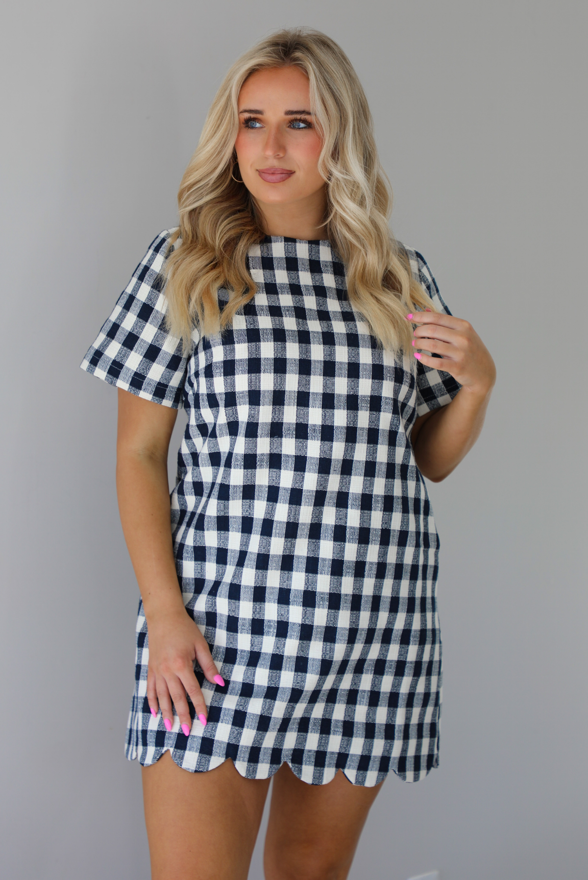 Woman wearing a dress with black and white gingham print pattern throughout. The dress has short sleeves, a high neckline, relaxed fit design, functional pockets on either side, and a scalloped hem. The photo is taken in front of a white wall.
