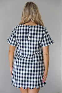 Woman wearing a dress with black and white gingham print pattern throughout. The dress has short sleeves, a high neckline, relaxed fit design, functional pockets on either side, and a scalloped hem. The photo shows the back of the dress and a closeup of the button closure trio in the back. 