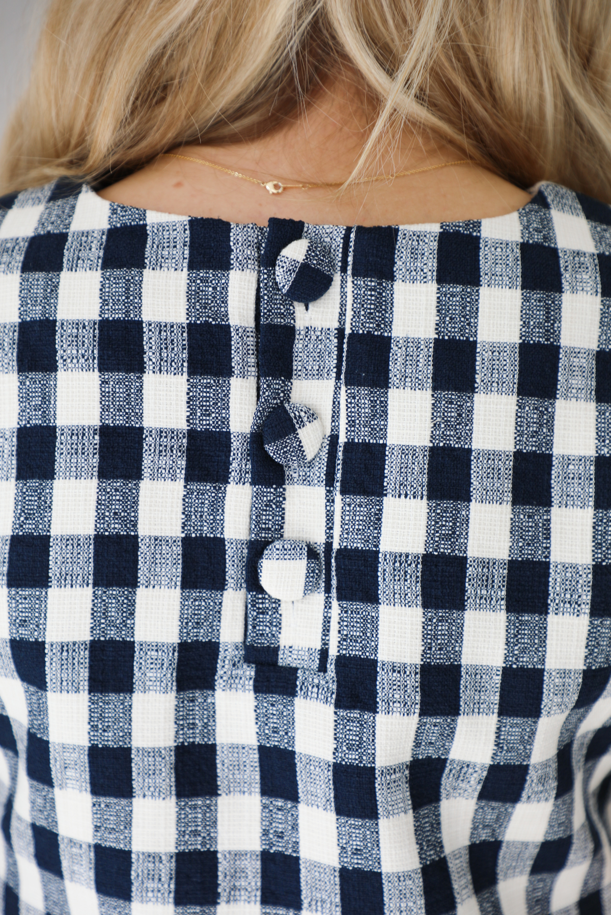 Woman wearing a dress with black and white gingham print pattern throughout. The dress has short sleeves, a high neckline, relaxed fit design, functional pockets on either side, and a scalloped hem. The photo shows a closeup of the button closure trio in the back. 