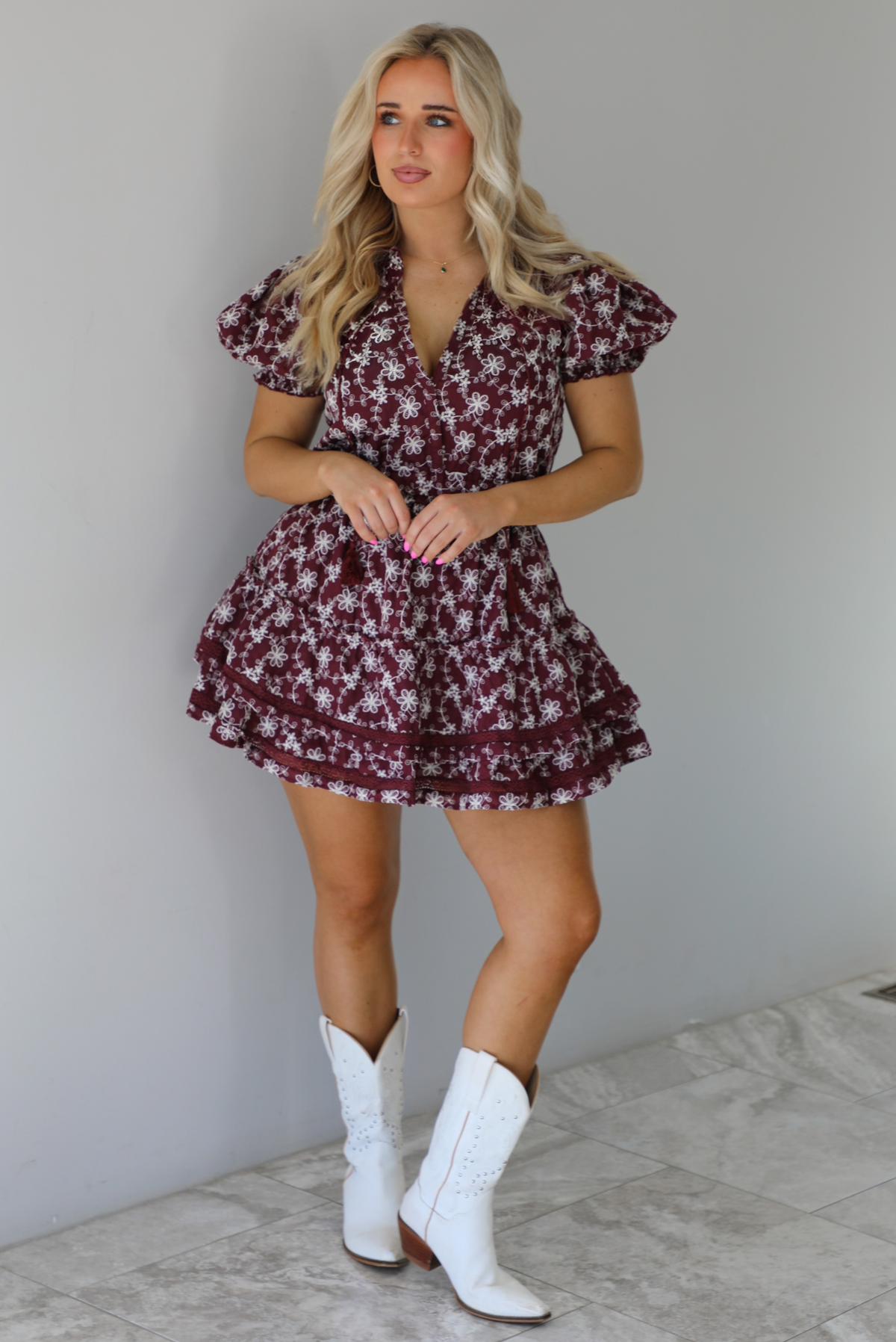 Woman wearing a wine dress that has an embroidered white floral print pattern, v-neckline, puffed sleeves, cinched with elastic waistline, and puffed hem skirt. The dress is paired with white cowboy boots. The photo is taken indoors against a white wall with white marble flooring.