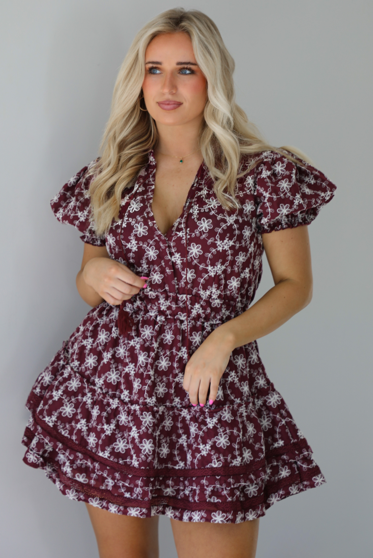 Woman wearing a wine dress that has an embroidered white floral print pattern, v-neckline, puffed sleeves, cinched with elastic waistline, and puffed hem skirt.  The photo is taken indoors against a white wall.