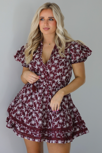 Woman wearing a wine dress that has an embroidered white floral print pattern, v-neckline, puffed sleeves, cinched with elastic waistline, and puffed hem skirt.  The photo is taken indoors against a white wall.