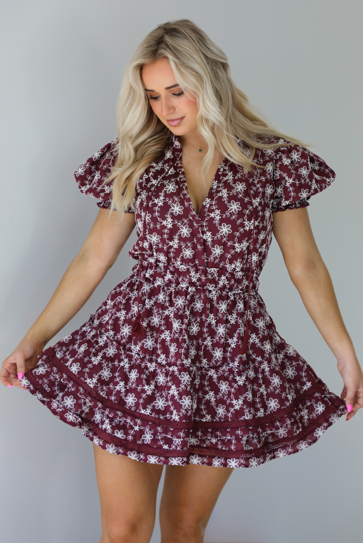 Woman wearing a wine dress that has an embroidered white floral print pattern, v-neckline, puffed sleeves, cinched with elastic waistline, and puffed hem skirt.  The photo is taken indoors against a white wall.
