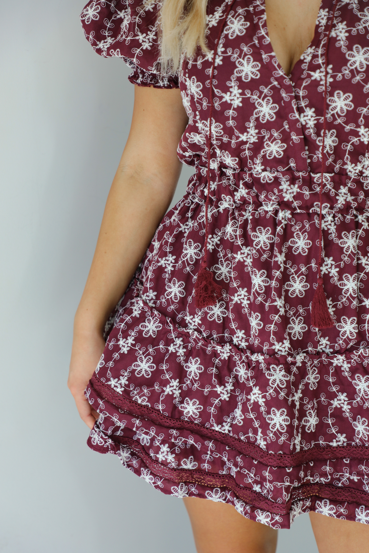 Woman wearing a wine dress that has an embroidered white floral print pattern, v-neckline, puffed sleeves, cinched with elastic waistline, and puffed hem skirt.  The photo is taken indoors against a white wall. The photo shows a closeup of the floral pattern of the dress, as well as the wine lace detailing at the hem. 