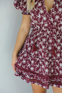 Woman wearing a wine dress that has an embroidered white floral print pattern, v-neckline, puffed sleeves, cinched with elastic waistline, and puffed hem skirt.  The photo is taken indoors against a white wall. The photo shows a closeup of the floral pattern of the dress, as well as the wine lace detailing at the hem. 
