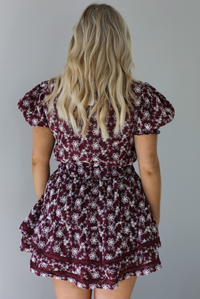 Woman wearing a wine dress that has an embroidered white floral print pattern, v-neckline, puffed sleeves, cinched with elastic waistline, and puffed hem skirt.  The photo is taken indoors against a white wall. The photo shows the back of the dress. 