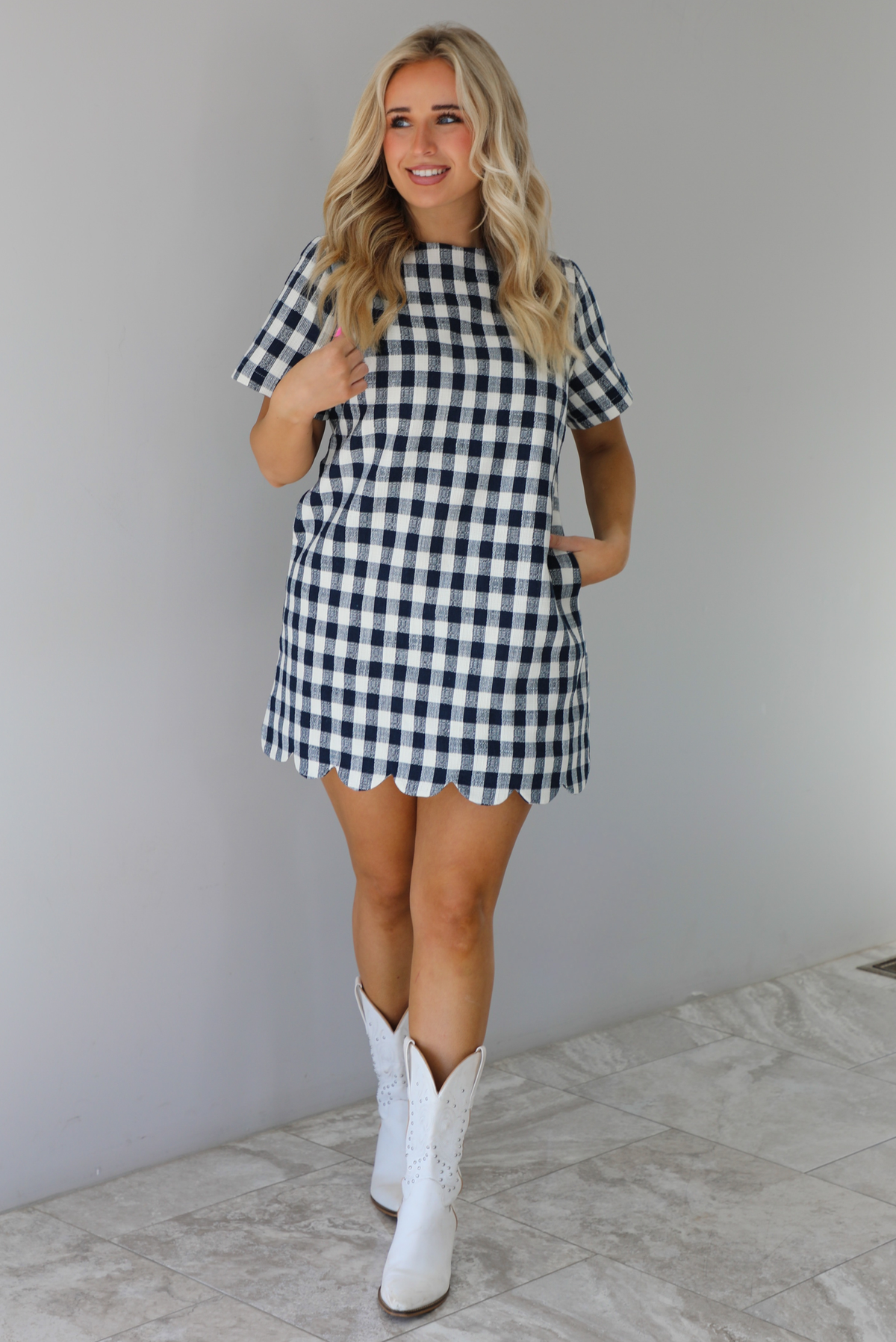Woman wearing a dress with black and white gingham print pattern throughout. The dress has short sleeves, a high neckline, relaxed fit design, functional pockets on either side, and a scalloped hem. The dress is paired with white cowboy boots. The photo is taken in front of a white wall with white marble flooring. 