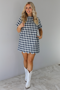 Woman wearing a dress with black and white gingham print pattern throughout. The dress has short sleeves, a high neckline, relaxed fit design, functional pockets on either side, and a scalloped hem. The dress is paired with white cowboy boots. The photo is taken in front of a white wall with white marble flooring. 