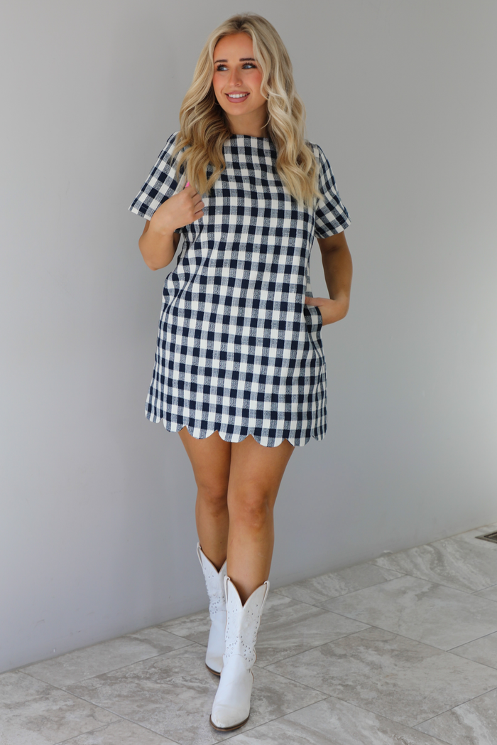 Woman wearing a dress with black and white gingham print pattern throughout. The dress has short sleeves, a high neckline, relaxed fit design, functional pockets on either side, and a scalloped hem. The dress is paired with white cowboy boots. The photo is taken in front of a white wall with white marble flooring. 