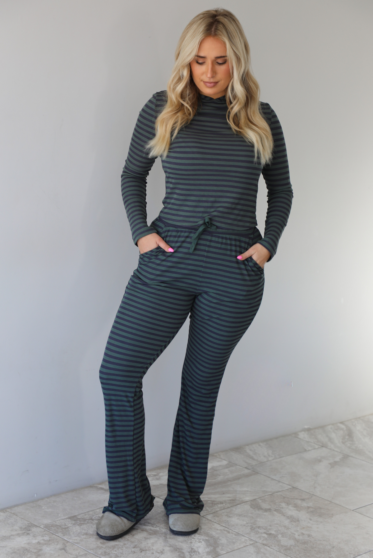 Woman wearing a navy and olive striped patterned set featuring a hoodie top and high waisted pants. The photo is taken indoors against a white wall with white marble flooring. 