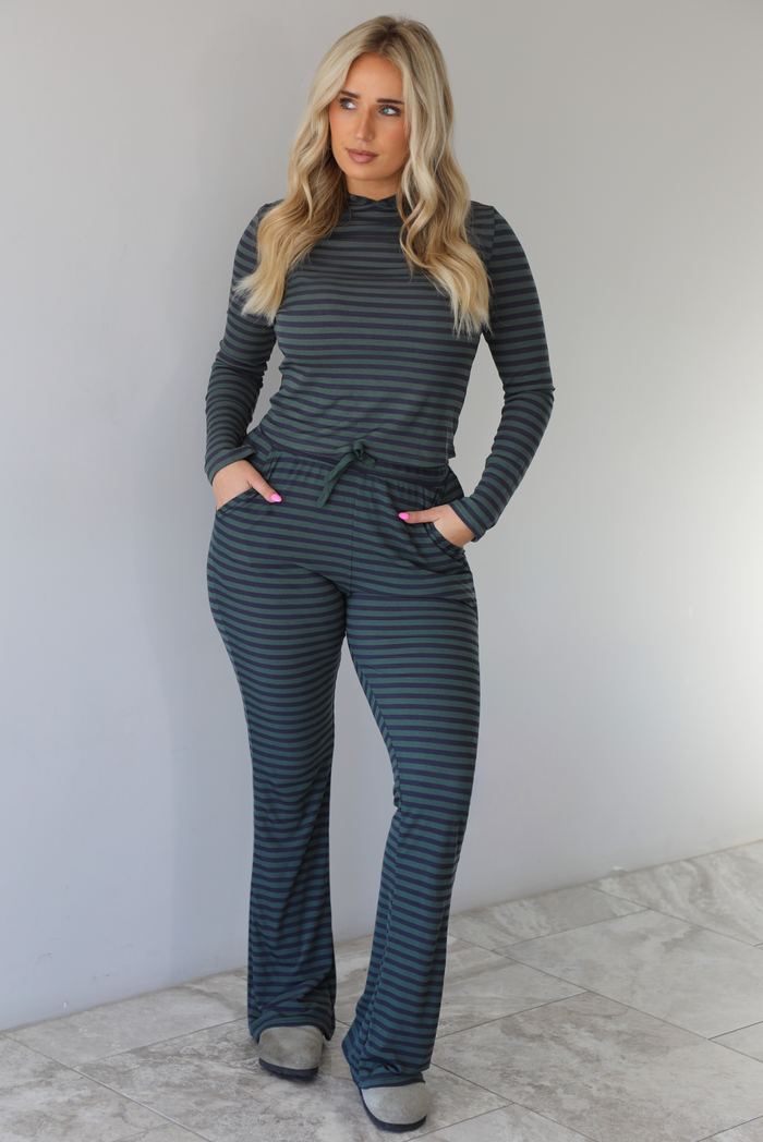 Woman wearing a navy and olive striped patterned set featuring a hoodie top and high waisted pants. The photo is taken indoors against a white wall with white marble flooring. 