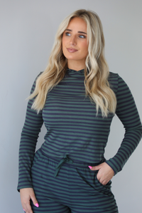 Woman wearing a navy and olive striped patterned set featuring a hoodie top and high waisted pants. The photo is taken indoors against a white wall. The photo shows a closeup of the hoodie. 