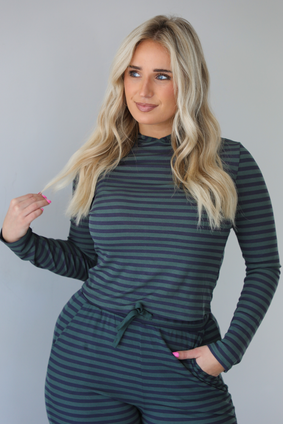Woman wearing a navy and olive striped patterned set featuring a hoodie top and high waisted pants. The photo is taken indoors against a white wall. The photo shows a closeup of the hoodie. 