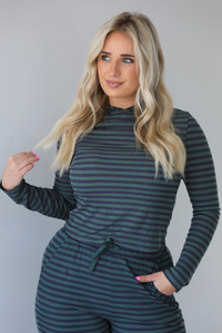 Woman wearing a navy and olive striped patterned set featuring a hoodie top and high waisted pants. The photo is taken indoors against a white wall. The photo shows a closeup of the hoodie. 
