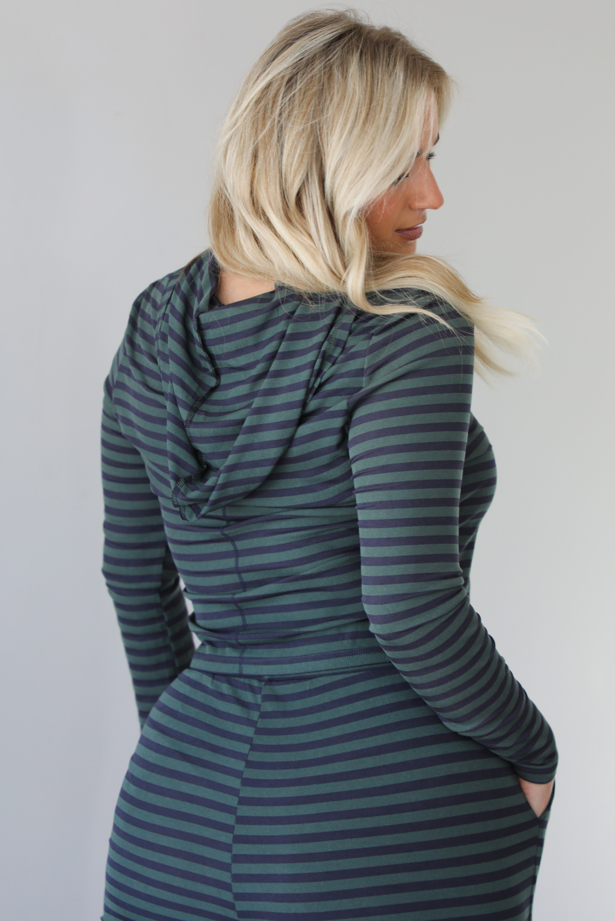 Woman wearing a navy and olive striped patterned set featuring a hoodie top and high waisted pants. The photo is taken indoors against a white wall. The photo shows a closeup of the back of the hoodie. 