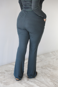 Woman wearing a navy and olive striped patterned set featuring a hoodie top and high waisted pants. The photo is taken indoors against a white wall with white marble flooring. The photo shows the back of the pants. 