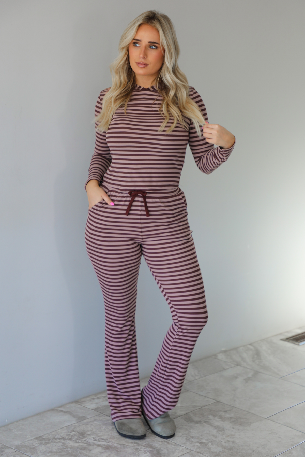 Woman wearing a burgundy and mauve striped patterned set featuring a hoodie top and high waisted pants. The photo is taken indoors against a white wall with white marble flooring. 