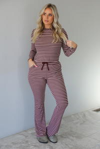 Woman wearing a burgundy and mauve striped patterned set featuring a hoodie top and high waisted pants. The photo is taken indoors against a white wall with white marble flooring. 