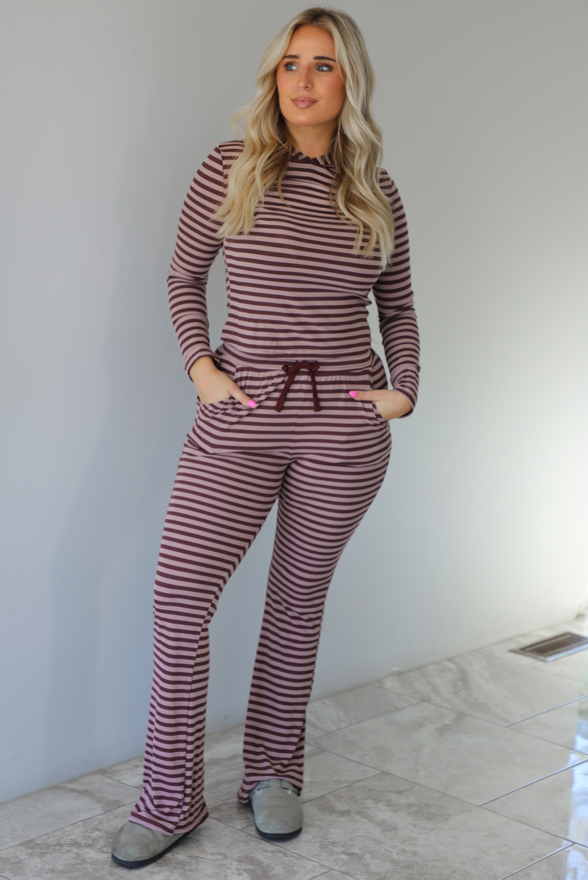 Woman wearing a burgundy and mauve striped patterned set featuring a hoodie top and high waisted pants. The photo is taken indoors against a white wall with white marble flooring. 