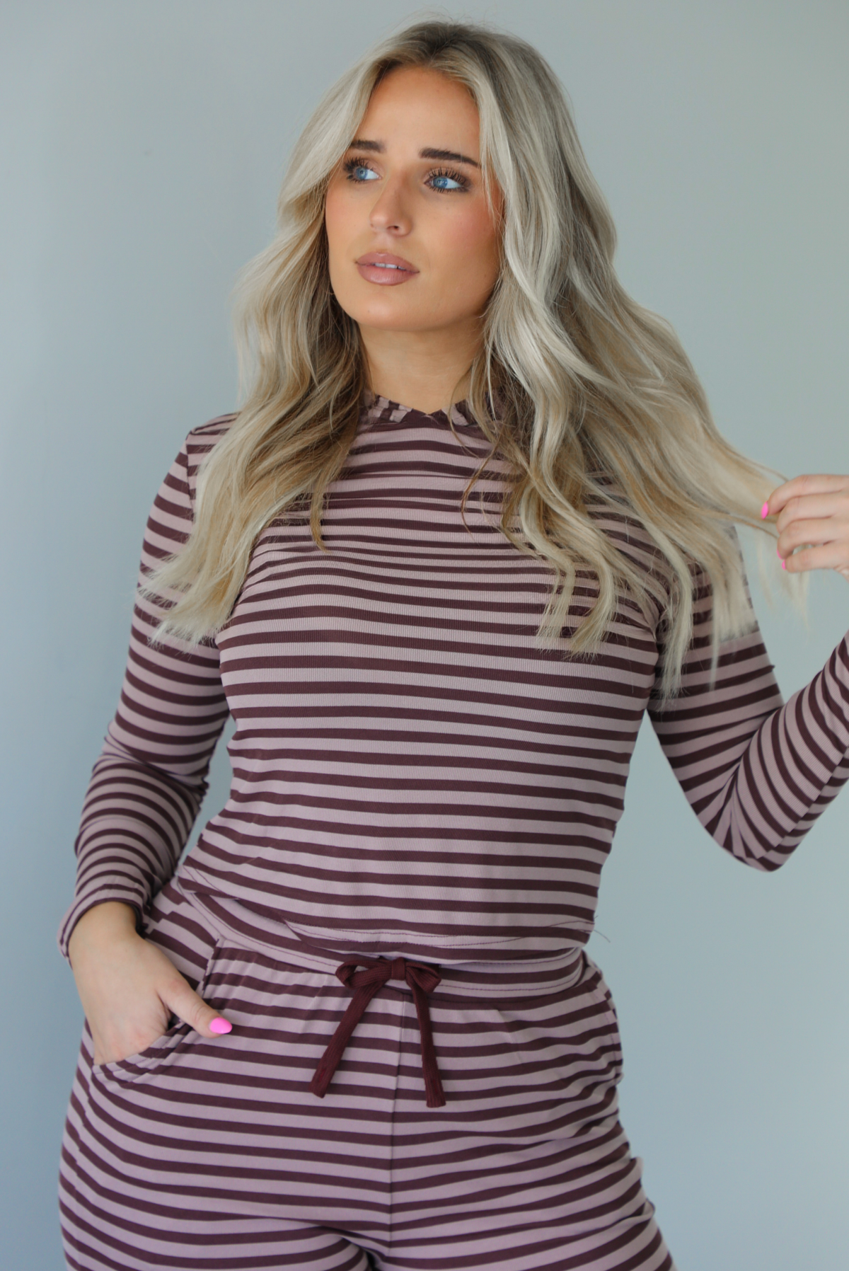 Woman wearing a burgundy and mauve striped patterned set featuring a hoodie top and high waisted pants. The photo is taken indoors against a white wall.